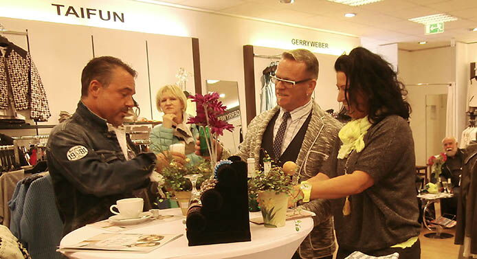 Spendenaktion im "Modehaus Fischer", wobei im Vordergrund Verantwortliche und Gäste an einem dekorierten Stehtisch direkt im Verkaufsraum des Modehauses stehen. Vertreten wird das Modehaus von der Filialleiterin Frau Vogel.