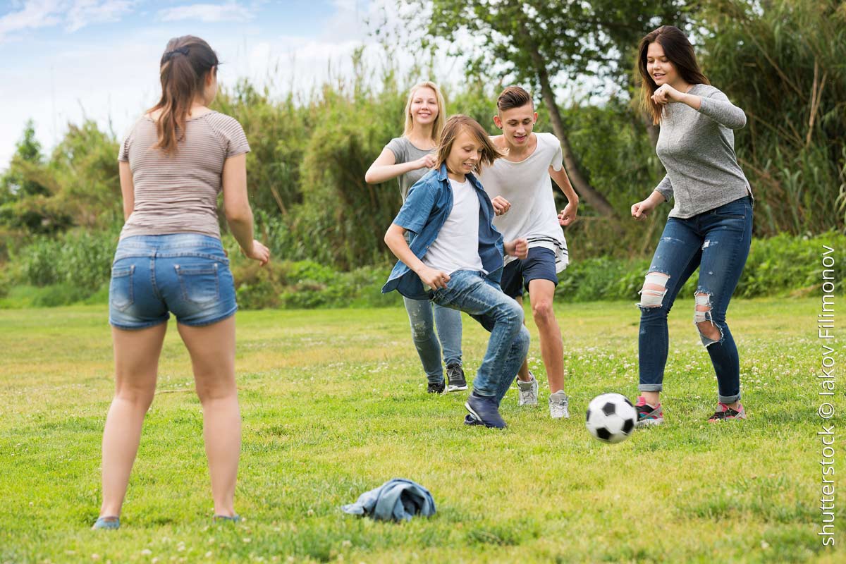 Fünf Jugendlichen, zwei Jungen und drei Mädchen zwischen 12 bis 16 Jahren, spielen auf dem grünem Rasen mit dem Fußball. Im Hintergrund sind Büsche und Bäume zu sehen. Die Kinder sind alle sommerlich und locker angezogen, tragen Jeans, Schorts und T-Shirt